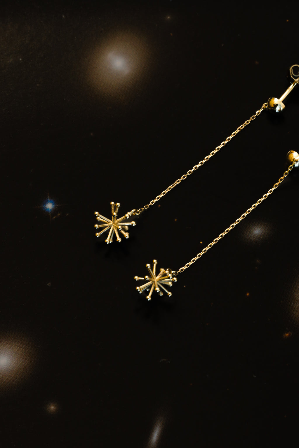 A pair of gold-toned dangle earrings with delicate chain straps and starburst-shaped charms, displayed against a dark, starry background.