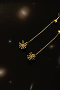 A pair of gold-toned dangle earrings with delicate chain straps and starburst-shaped charms, displayed against a dark, starry background.